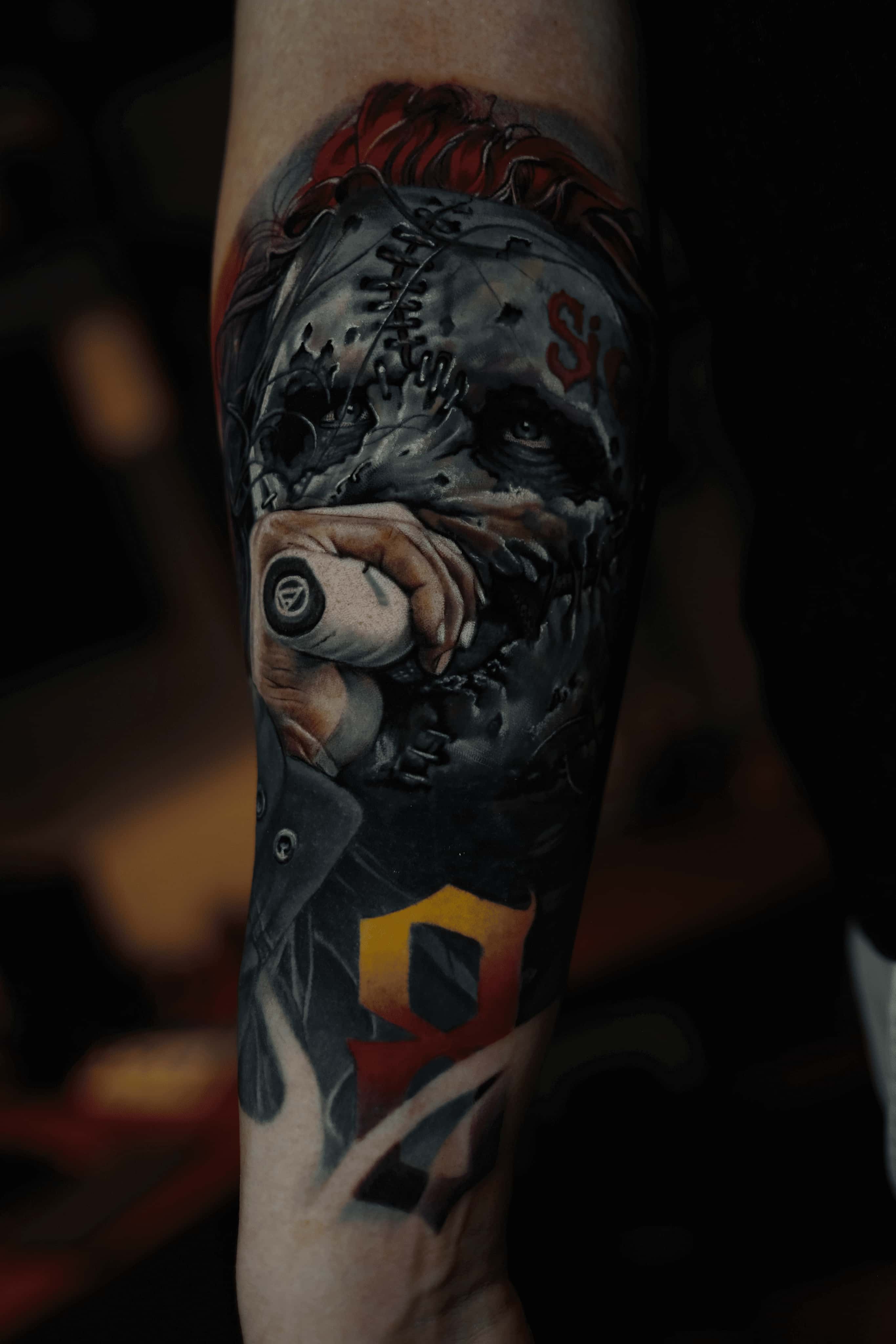 Tetovaža na podlaktici koja prikazuje detaljno lice sa ušivenom maskom frontmena benda Slipknot koji drži mikrofon, sa crvenom kosom i stilizovanim crveno-žutim brojem 9 ispod.
